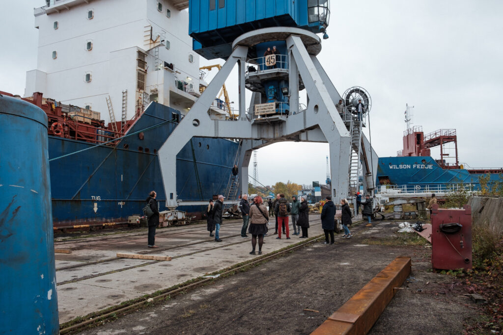 Open House Tallinn tuur Hundipeal külas. Grupp inimesi jalutab suure sadamakraana alt läbi, vasakul pool on sadamas suur kaubalaev.